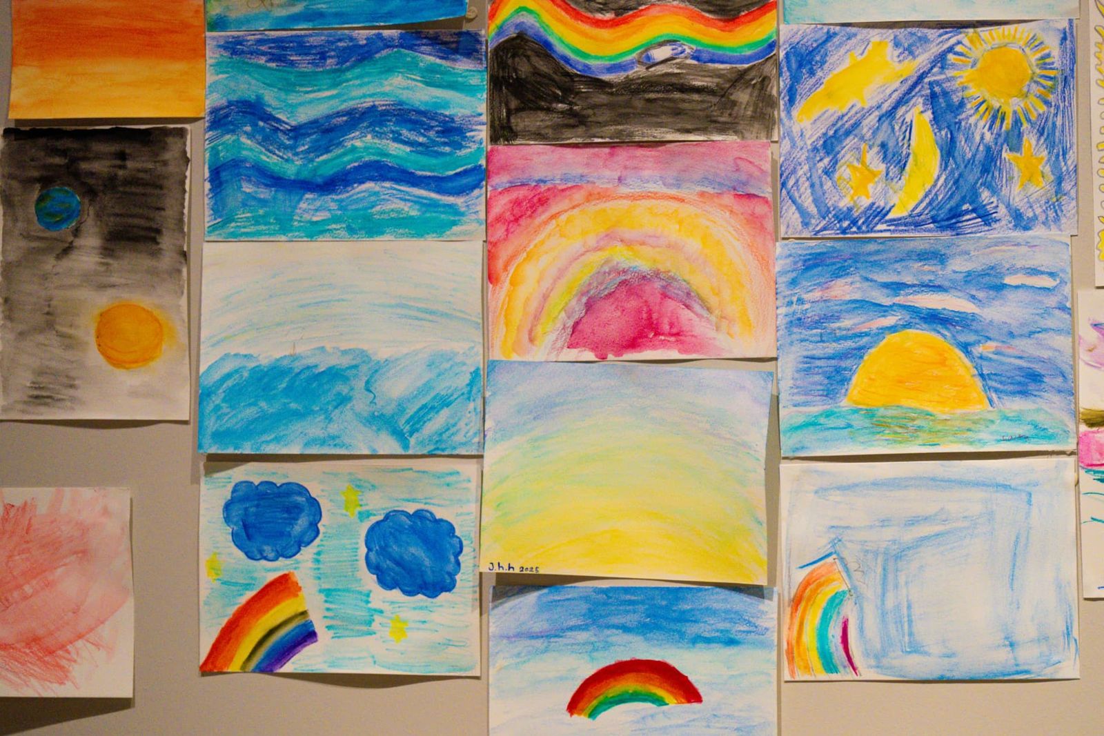 A wall of children's watercolour paintings of skies, suns and rainbows