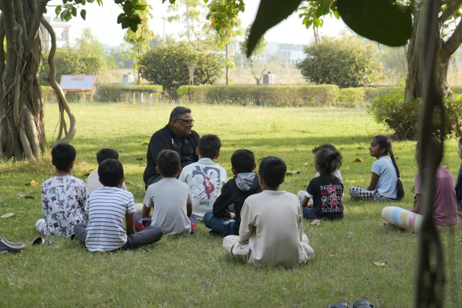 A teacher leading an outdoor lesson with children seated on the grass beneath a tree
