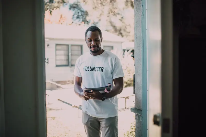 A volunteer in a Volunteer T-shirt taking notes on a doorstep during a household visit
